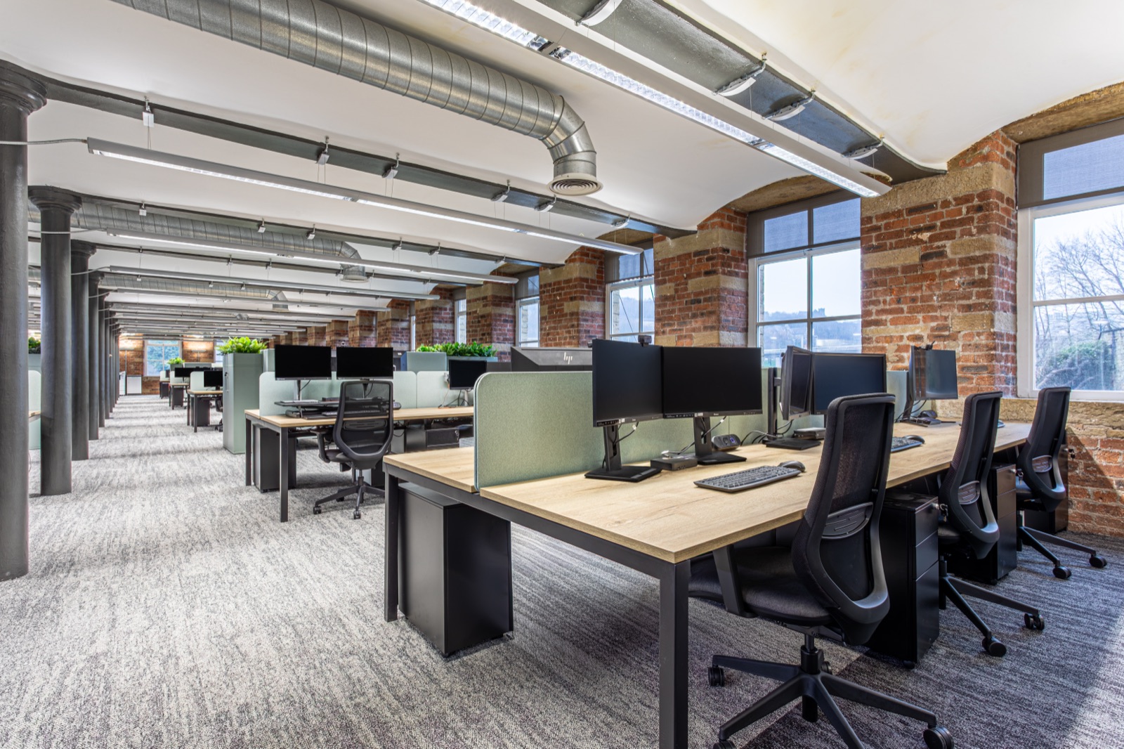 Appleyard Lees open-plan office in a converted mill, oak desks with green screens between cast-iron columns, beneath brick vaults and exposed ductwork