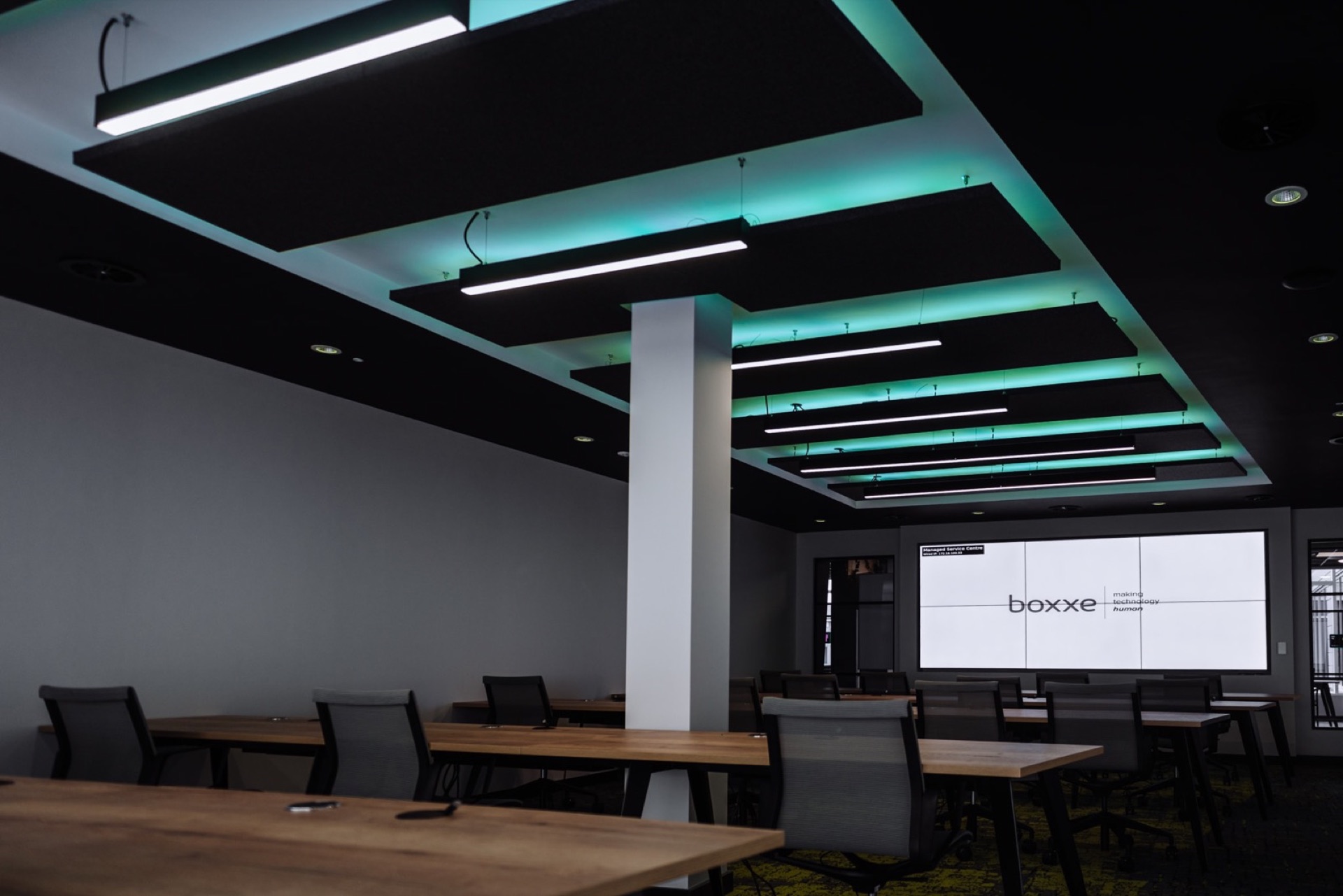 Presentation room at Boxxe, with rows of mesh-back chairs at long oak tables facing a four-panel video wall, beneath suspended acoustic rafts lit teal