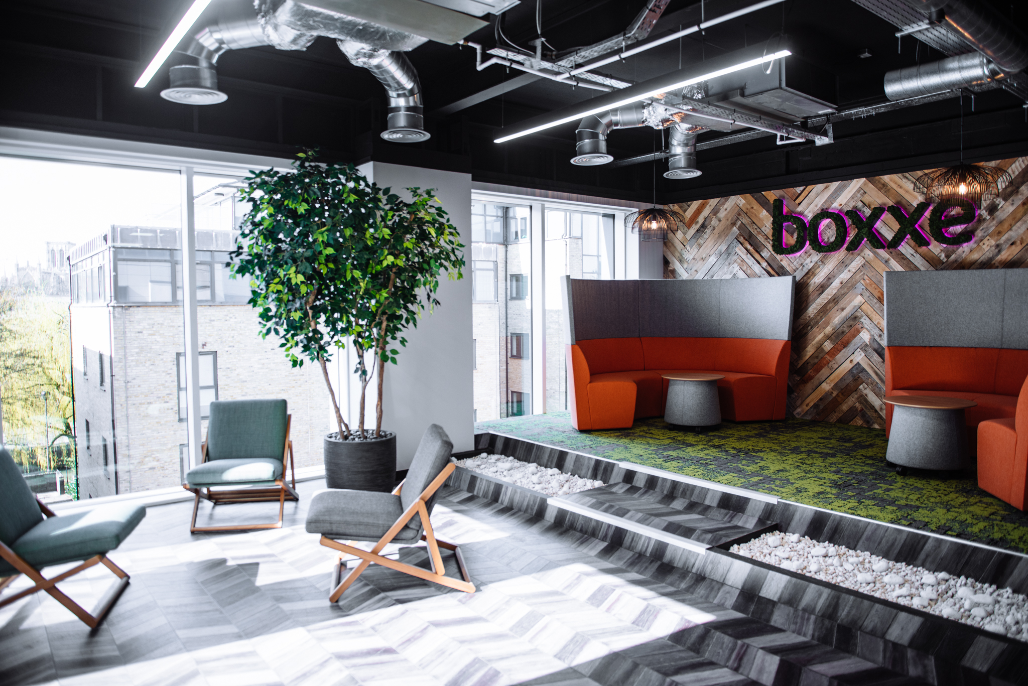 Curved orange booth pods with grey acoustic backs at Boxxe, on a raised green carpeted platform against a herringbone reclaimed-timber wall carrying the boxxe logo in moss