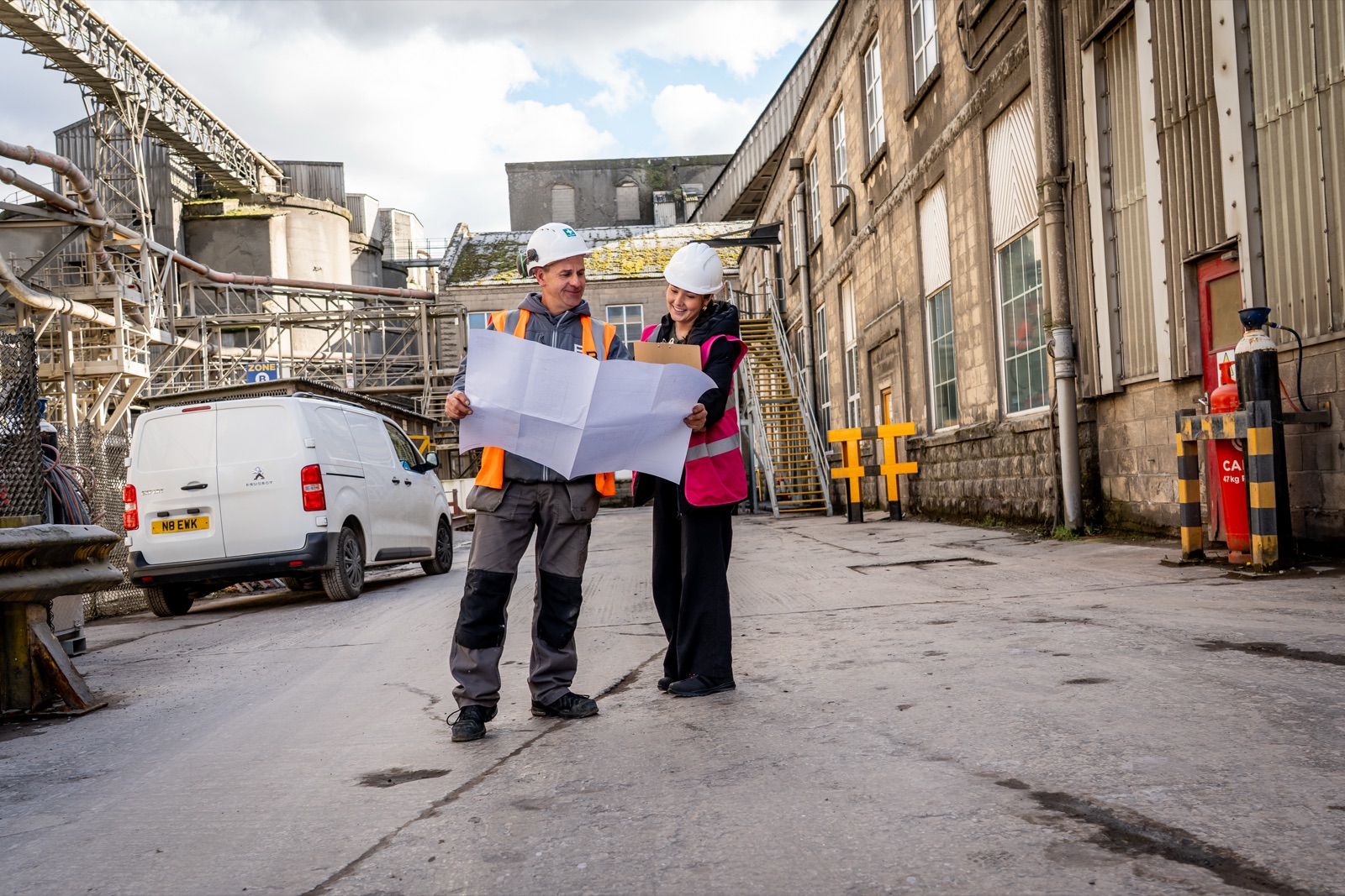 Two Building Interiors team members in hi-vis reviewing site drawings in the yard at Breedon, with cement silos behind them