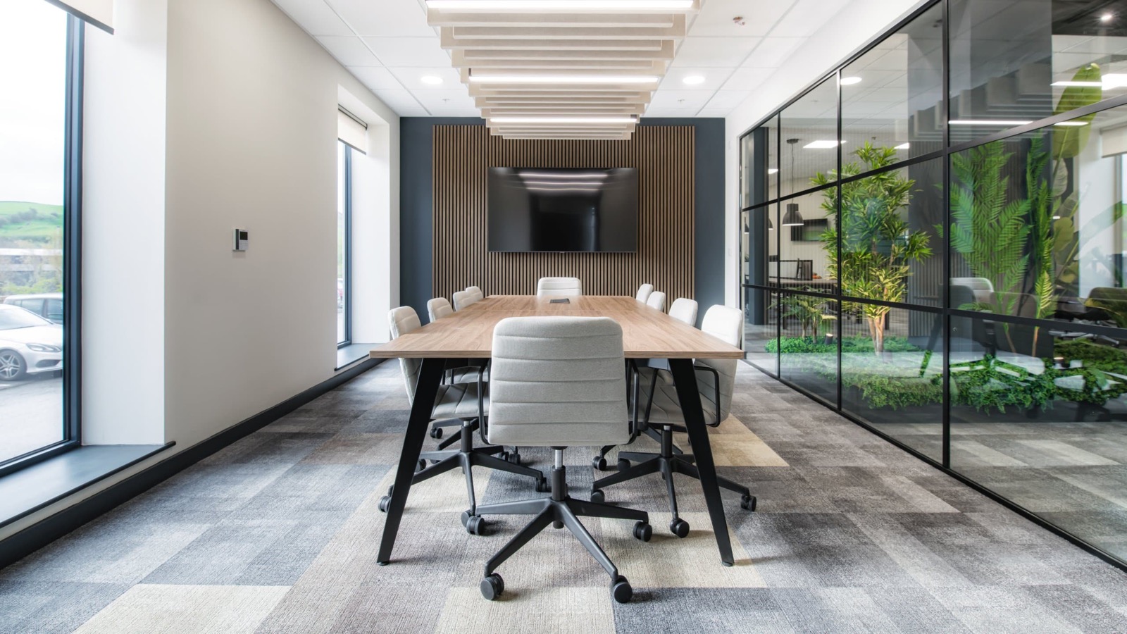 Meeting room at Green & Brown, a boardroom table beneath acoustic ceiling baffles, divided from the office beyond by a black-framed glazed partition with planting behind it