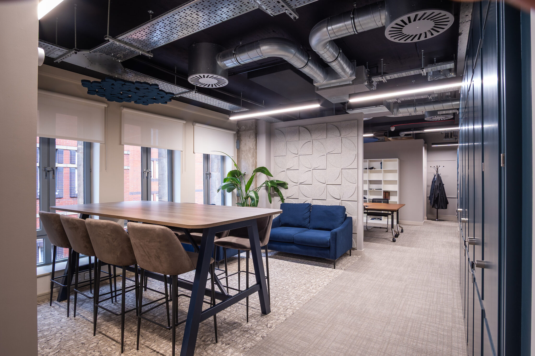 Project zone at Spire, a long timber table and chairs beside a blue sofa, beneath exposed ductwork and an acoustic feature wall