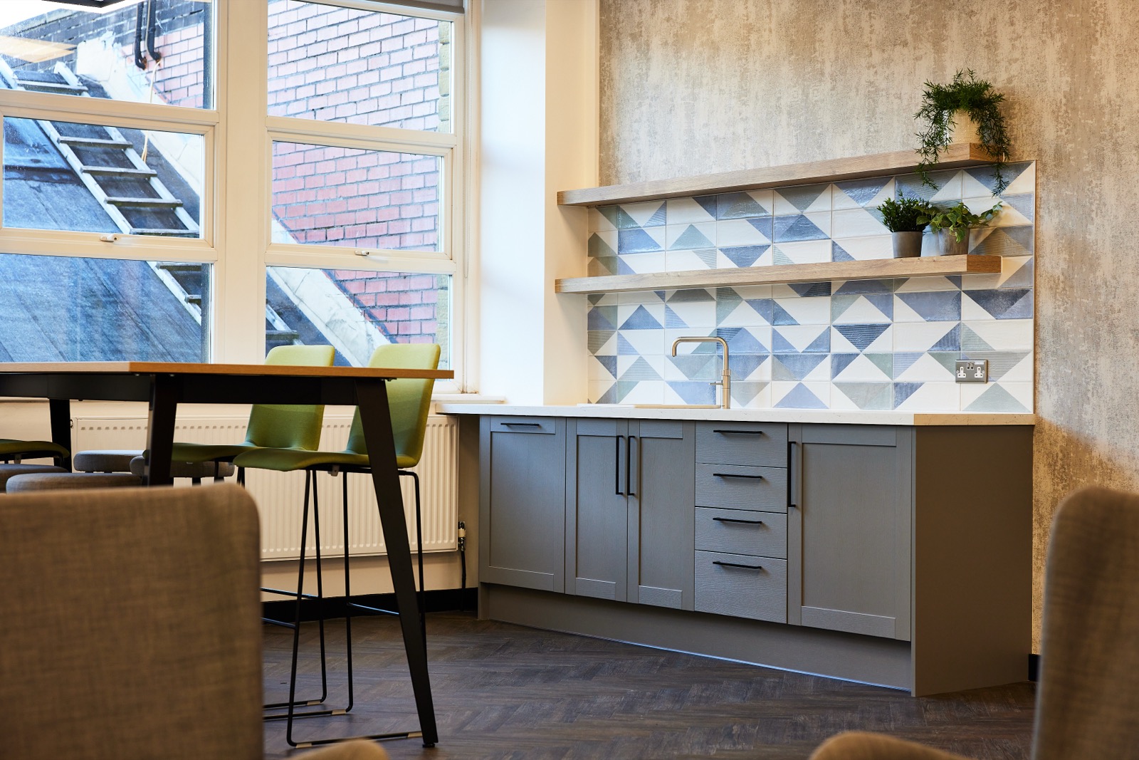 Tea point at Thornton & Ross, with grey shaker units, a brass tap and a geometric tiled splashback, beside a poseur table with green chairs